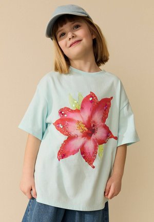 Junges Kind trägt eine hellblaue Mütze und ein übergroßes hellblaues T-Shirt mit großem roten und pinkfarbenem Blumenprint, verziert mit Pailletten.