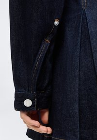 Χέρι που φοράει μανίκι σκούρου μπλε τζιν μπουφάν με πορτοκαλί ραφές και ασημένιο κουμπί κοντά στον καρπό, σε απλό φόντο.