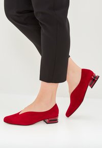 Rote Wildleder-Absatzschuhe mit niedrigem Absatz, v-förmigem Design und quadratischem Absatz, versehen mit schwarzen und roten Akzenten. Getragen mit schwarzen, schmal zulaufenden Hosen.