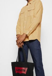 Chemise jaune à boutons avec deux poches poitrine, associée à un jean bleu foncé. Un sac fourre-tout noir avec un logo Levi's rouge est tenu devant.
