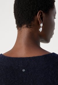 Marineblauwe gebreide trui met een ronde halslijn; beschikt over een klein metalen label. Model draagt oversized zilveren oorbellen, die de gladde textuur benadrukken.