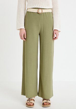 Kobieta ubrana w oliwkowe, szerokie spodnie z wysokim stanem, przewiązane plecionym beżowym paskiem, kremowy sweter z dzianiny oraz beżowe sandały z klamrami.