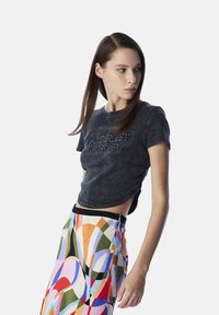 Schwarzes, cropped T-Shirt mit geprägtem Text; kombiniert mit einem bunten, gemusterten Rock, der Kurven in Pink, Orange, Blau, Grün und schwarzen Akzenten zeigt.
