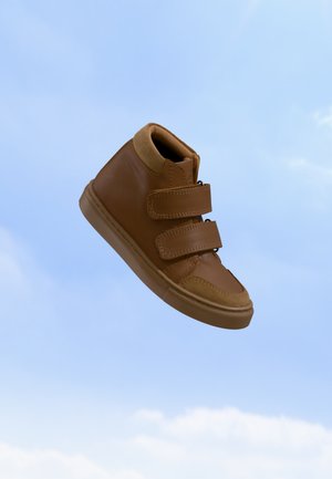 Baskets montantes marron avec deux bandes velcro flottant devant un ciel bleu clair.