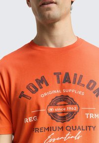 Pomarańczowa bawełniana koszulka z okrągłym dekoltem i krótkimi rękawami. Na przodzie znajduje się napis: "TOM TAILOR" w pogrubionej czerni, z okrągłymi akcentami logo.