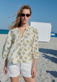 Blusa con stampa floreale con maniche a palloncino in giallo e bianco, con scollo a V. Abbinata a pantaloncini bianchi, ambientata su uno sfondo di spiaggia.