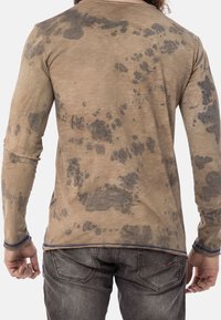 Camicia beige a maniche lunghe con motivo a macchie scure, caratterizzata da un collo rotondo e dettagli in cucitura blu lungo l'orlo e le maniche.
