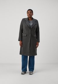 Prugasta siva jakna z dolgimi rokavi, dvema sprednjima žepoma in gumbnim zapiranjem, nošena čez sivo majico, v kombinaciji z modrimi širokimi jeans hlačami in supergami.