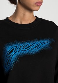 Primer plano de una persona que lleva una camiseta negra con un gráfico de puntos azules y el logo "Guess" en el pecho, mostrando el cuello, los labios y un pendiente.