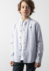 Jasnoniebieska, teksturowana koszula z długim rękawem, z kołnierzykiem typu mandarynka, drewnianymi guzikami oraz małym granatowym logo na lewej piersi. Nosi się ją na białą koszulę.