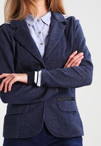 Marinblå blazer i texturerad tyg med skräddarsydd passform. Har två framfickor, kontrasterande vit och svart manschettsträngar, samt spetsiga slag.