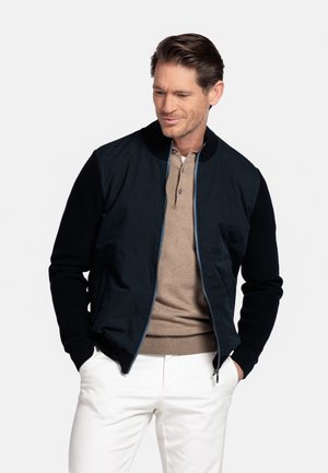 Man met een marineblauwe jas met rits over een beige henley-shirt en witte broek, handen in de zakken, kijkt omlaag met een lichte glimlach.