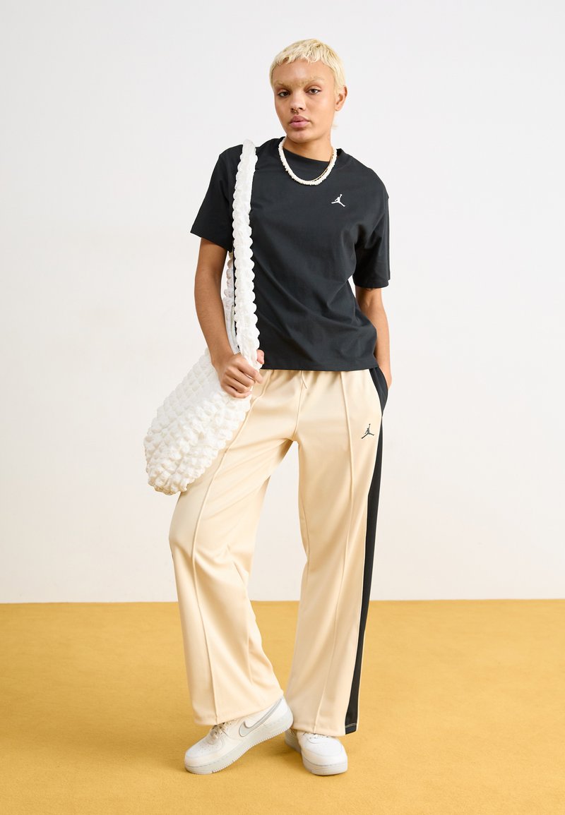 Schwarzes T-Shirt mit kurzen Ärmeln von Jordan, beiger Jogginghose mit schwarzem Seitenstreifen, weiße strukturierte Tasche. Modell trägt weiße Sneakers.