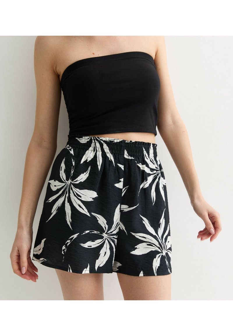 Femme portant un top bandeau noir et un short noir avec un grand motif de feuille tropicale blanche, debout devant un fond blanc uni.