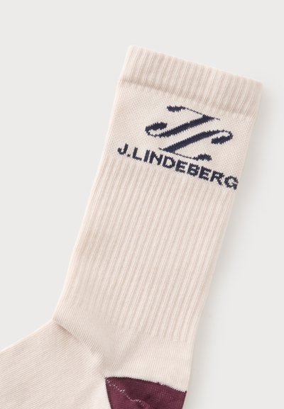 Chaussette côtelée couleur crème avec talon violet foncé et logo noir J. Lindeberg ainsi qu’un texte près du haut du bord-côte.