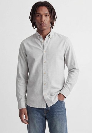 Homme avec des dreadlocks de longueur moyenne portant une chemise gris clair à boutons et un jean bleu, debout avec une main dans la poche devant un fond uni.