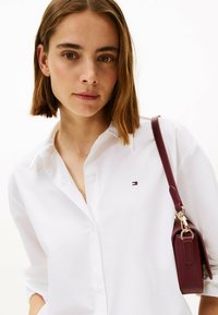 Camisa blanca de botones con un sutil detalle de logo, combinada con un pequeño bolso burdeos que presenta un acento de hardware dorado.