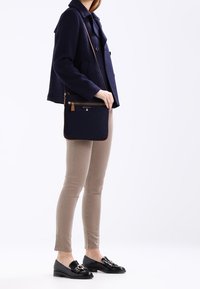 Borsa a tracolla blu navy con una texture liscia, chiusura con zip, dettagli in pelle e un design sottile, indossata da un modello con giacca blu navy e pantaloni beige.