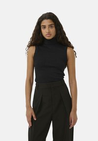 Schwarzes ärmelloses Rollkragensweatshirt in Rippstruktur, kombiniert mit hochtaillierten, maßgeschneiderten schwarzen Hosen mit Bundfalten. Einfaches, monochromatisches Design.