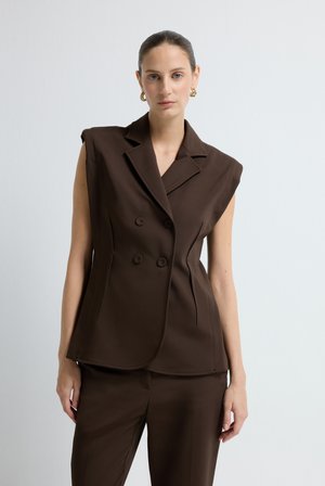 Donna con capelli raccolti indossante un blazer doppiopetto marrone senza maniche e pantaloni abbinati, in piedi contro uno sfondo bianco uniforme.