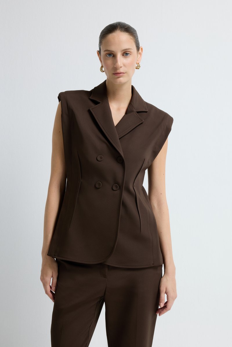 Frau mit zurückgegeltem Haar, die ein ärmelloses braunes, doppelt knöpfiges Blazer und passende Hose trägt, steht vor einem schlichten weißen Hintergrund.