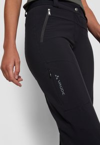 Black technical pants with a slim fit, featuring a zippered pocket, flat seams, and a logo on the lower leg; made from stretchy, durable fabric.