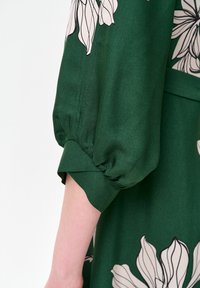 Zielona sukienka w kwiaty z długimi, bufkami; jedwabista tekstura, z dużymi wzorami kremowych kwiatów; dopasowana przy mankietach.