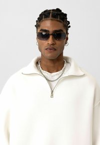 Witte zip-up sweatshirt met een hoge kraag, gelaagd over een wit t-shirt. Voorzien van zilveren kettingen en donkere zonnebril. Gladde textuur.