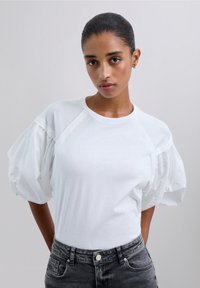 Camiseta blanca con mangas abullonadas hecha de un material transparente, con un escote redondo y un cuerpo ajustado, combinada con jeans gris oscuro.
