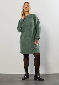 Abito in maglia verde con una vestibilità comoda, scollo rotondo e maniche lunghe. Abbinato a stivali neri alla caviglia e collant opachi.