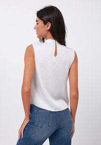White sleeveless top with a high collar and button closure at the back; smooth texture, complemented by blue denim jeans.
