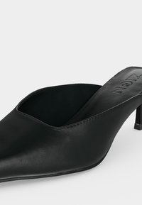 Mule en cuir noir à bout pointu avec un talon stiletto bas et un design slip-on, marqué "ALCEN" sur la semelle intérieure.