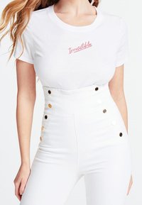 Camiseta blanca con "Irresistible" en un script rosa, combinada con jeans blancos de cintura alta que tienen acentos de botones dorados y negros a lo largo del costado.