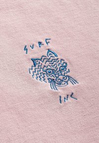 Różowy materiał o teksturowanej powierzchni z niebieskim haftem przedstawiającym stylizowaną twarz kota oraz słowami "SURF INC" poniżej.