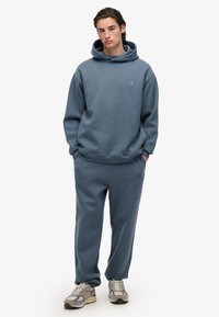 Felpa blu con cappuccio e pantaloni abbinati in tessuto morbido. La felpa presenta una grande tasca frontale e polsini a coste. Sneakers nei toni del grigio.
