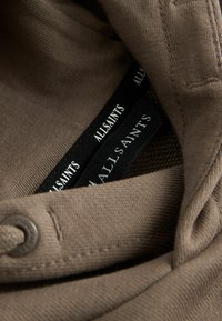 Tissu marron avec une finition texturée, comportant une fermeture à bouton et des étiquettes noires avec un texte blanc indiquant "ALLSAINTS".