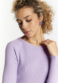 Maglione lilla a coste con scollatura rotonda. La donna indossa orecchini circolari e un anello viola abbinati. La morbida texture e il design aderente mettono in risalto la forma.