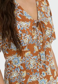 Bruine bloemjurk met een diepe V-hals, een klein strikdetail en intrieste patronen in blauw en geel, van zachte stof en met korte mouwen.