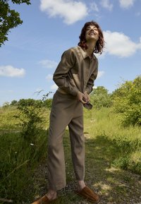 Jeune personne portant une chemise et un pantalon beige debout sur un chemin herbeux, souriant sous un ciel bleu partiellement nuageux.