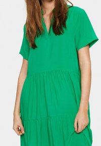 Robe en coton verte avec une coupe courte et décontractée, col en V, manches courtes et jupes à volants froncés. Texture lisse et légère.