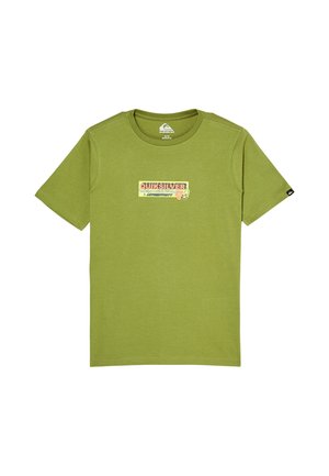 Jongen met oranje muts, groene Quiksilver T-shirt, losse blauwe spijkerbroek met opgerolde pijpen en zwarte sneakers, staat en lacht.