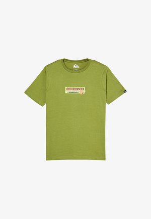 Chłopiec w pomarańczowej czapce, zielonym t-shircie Quiksilver, luźnych niebieskich dżinsach podwiniętych u dołu i czarnych trampkach, stoi i się uśmiecha.