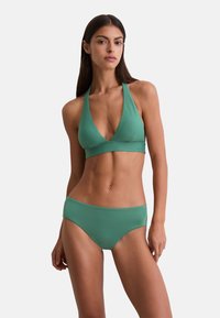 Mujer con cabello largo y oscuro que lleva un top de bikini verde tipo halter y una parte inferior a juego, de pie contra un fondo claro y liso.