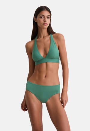 Vrouw met lang, donker haar in een groene halter bikini-top en bijpassende bikini-broek, staande tegen een effen, lichte achtergrond.