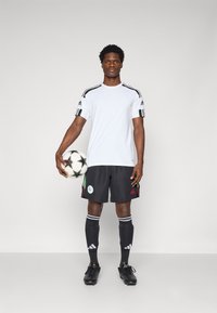 Bílé sportovní tričko s černými pruhy, černé šortky se zelenými akcenty, černé ponožky a černé boty, držící bílý fotbalový míč s černými hvězdami.