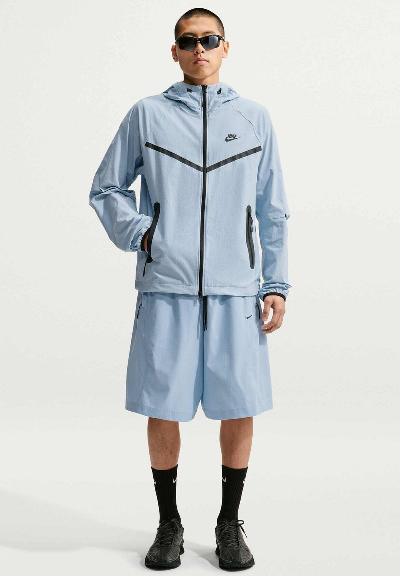 Junger Mann trägt eine hellblaue Nike-Windjacke und Shorts, schwarze Nike-Socken, schwarze Sneakers und dunkle Sonnenbrille, steht mit einer Hand in der Tasche.