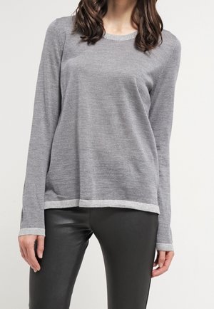 Kobieta ubrana w jasnoszary sweter z długim rękawem z kontrastującymi mankietami i krawędzią, zestawiony z czarnymi spodniami z ekologicznej skóry.