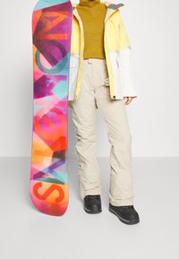 Snowboard mit einem bunten, verlaufenden Design, das mit fetten Buchstaben versehen ist, kombiniert mit einer gelben und weißen Jacke, beigen Hosen und schwarzen Stiefeln.