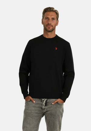 Schwarzer Sweatshirt aus Baumwolle, mit Rundhalsausschnitt, gerippten Bündchen und einem kleinen roten Logo auf der Brust. Getragen mit hellgrauen Jeans.