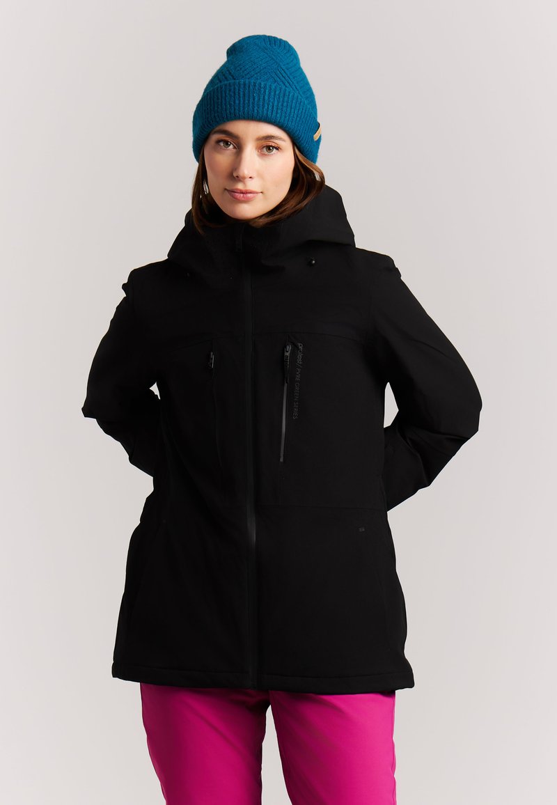 Femme portant une veste noire, un bonnet tricoté bleu et un pantalon rose, debout les mains dans le dos, devant un fond clair uni.